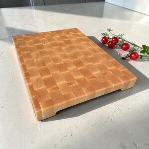 Maple end grain butcher block angled on a marble counter, showing its thickness and carved side handle grooves, with cherry tomatoes on the vine beside it.