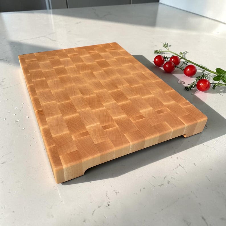 Maple end grain butcher block angled on a marble counter, showing its thickness and carved side handle grooves, with cherry tomatoes on the vine beside it.