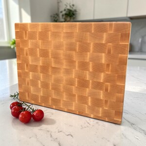 Maple end grain cutting board standing upright on marble, showing the full checkerboard face head-on, with cherry tomatoes and rosemary in front and a blurred kitchen behind.