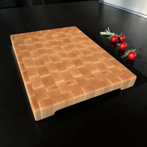 Maple end grain butcher block with support feet on a dark counter, rosemary and cherry tomatoes behind it
