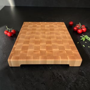 May include: A rectangular wooden cutting board featuring an end-grain construction with a woven-like pattern of light brown wood blocks. The board has a thick profile with small cutouts on the bottom edges and sits on a dark surface with tomatoes.