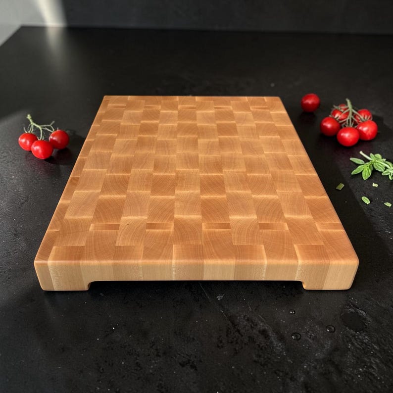 May include: A rectangular wooden cutting board featuring an end-grain construction with a woven-like pattern of light brown wood blocks. The board has a thick profile with small cutouts on the bottom edges and sits on a dark surface with tomatoes.