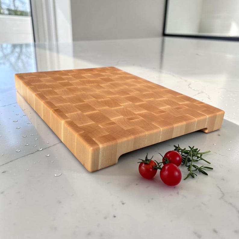 Maple end grain cutting board resting on a marble counter in a bright, window-lit kitchen, with cherry tomatoes and a rosemary sprig in front.