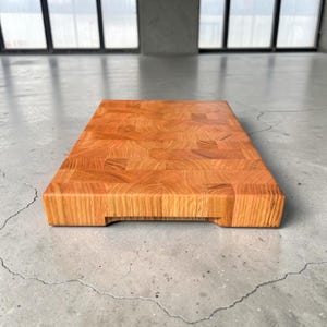 May include: A rectangular end-grain butcher block cutting board made of light brown wood. The board has a smooth surface and a thick profile, with a small recessed area on the bottom edge. The wood grain pattern is visible throughout.