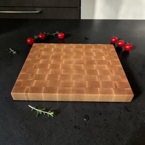 May include: A square wooden cutting board with an end-grain checkerboard pattern in light brown tones. The board is surrounded by several small red cherry tomatoes and small sprigs of green herbs, highlighting the natural wood grain texture.