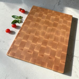 Maple end grain chopping block viewed at an angle on marble, with cherry tomatoes and fresh basil arranged at the upper corner.