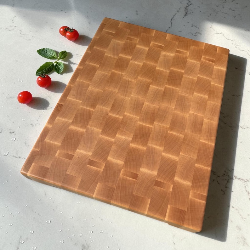 Maple end grain chopping block viewed at an angle on marble, with cherry tomatoes and fresh basil arranged at the upper corner.