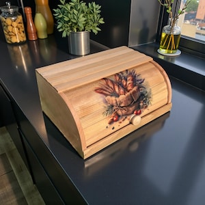 May include: Wooden bread box with a roll-top lid. The front features a painted design of a basket filled with various types of bread, berries, and lavender. The box has a light wood finish and a small wooden knob.