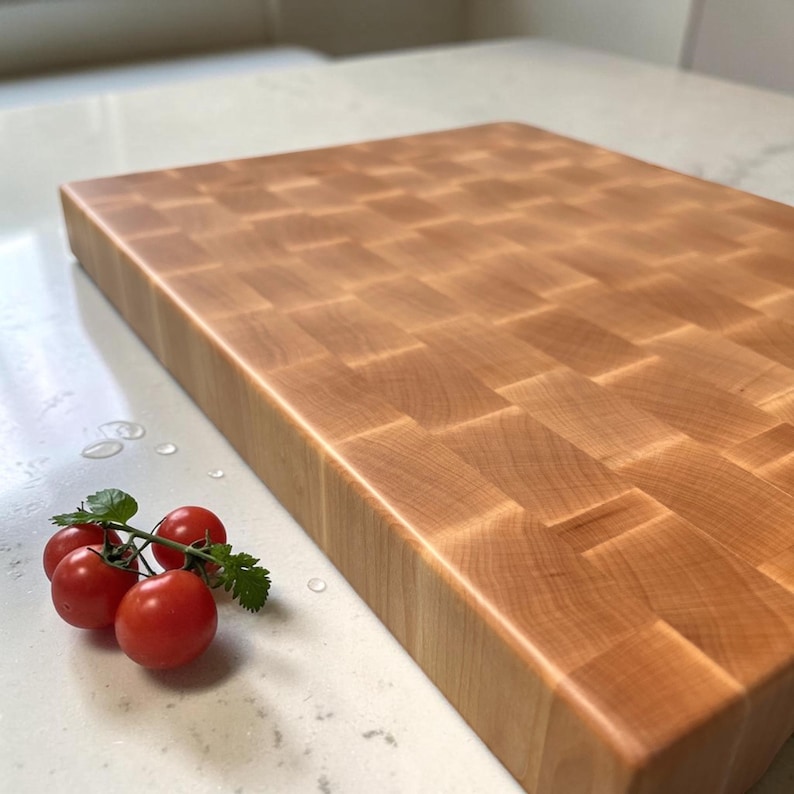 End grain maple cutting board photographed at an angle on a marble countertop, with a cluster of fresh cherry tomatoes on the vine in front.