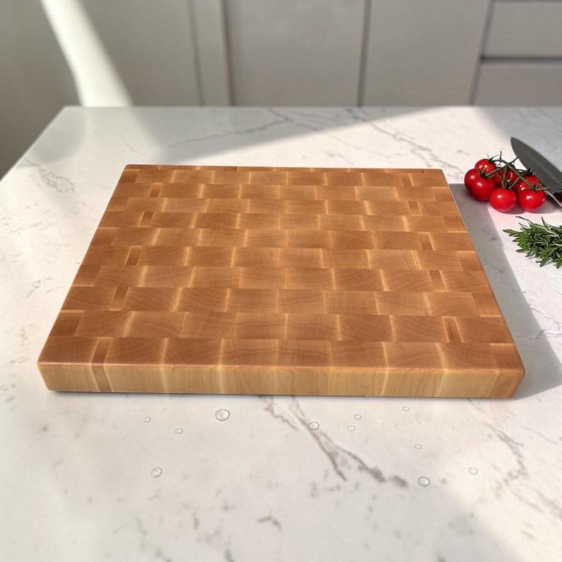Maple end grain cutting board lying at a low angle on a marble counter, with cherry tomatoes, rosemary and a knife at the top corner.