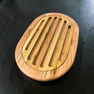 May include: Oval-shaped wooden breadboard with a grooved center for slicing. The board features a light brown and reddish-brown wood grain pattern. The outer rim is a lighter shade of wood, creating a contrasting border.