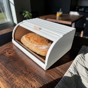 May include: A white wooden bread bin with a curved top and a light brown interior. A loaf of bread sits inside the bin. The bin is on a dark wooden table.