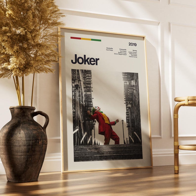 May include: A vertical film poster for the 2019 movie Joker, displaying the title in bold black text, production credits, and a central image of a person in a red suit dancing on outdoor stairs. The poster has a light background and a thin frame.