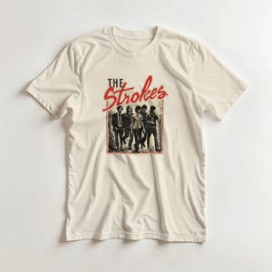 Könnte beinhalten: Cremefarbenes T-Shirt mit roter und schwarzer Grafik. Die Grafik zeigt den Bandnamen "The Strokes" in einer stilisierten Schriftart über einem Schwarz-Weiß-Bild der Bandmitglieder. Das T-Shirt hat kurze Ärmel und einen Rundhalsausschnitt.