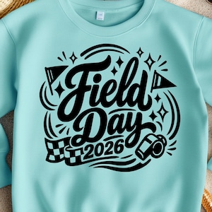May include: A light blue sweatshirt with the words "Field Day 2026" in a black, stylized font. The design includes flags, stars, and a checkered flag. The sweatshirt is surrounded by a straw hat, sunglasses, starfish, and sand.