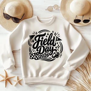 May include: Cream-colored sweatshirt with the words "Field Day 2026" in a black, decorative font. The design includes flags, stars, and a checkered flag. The sweatshirt is laid flat with sunglasses and a straw hat in the background.