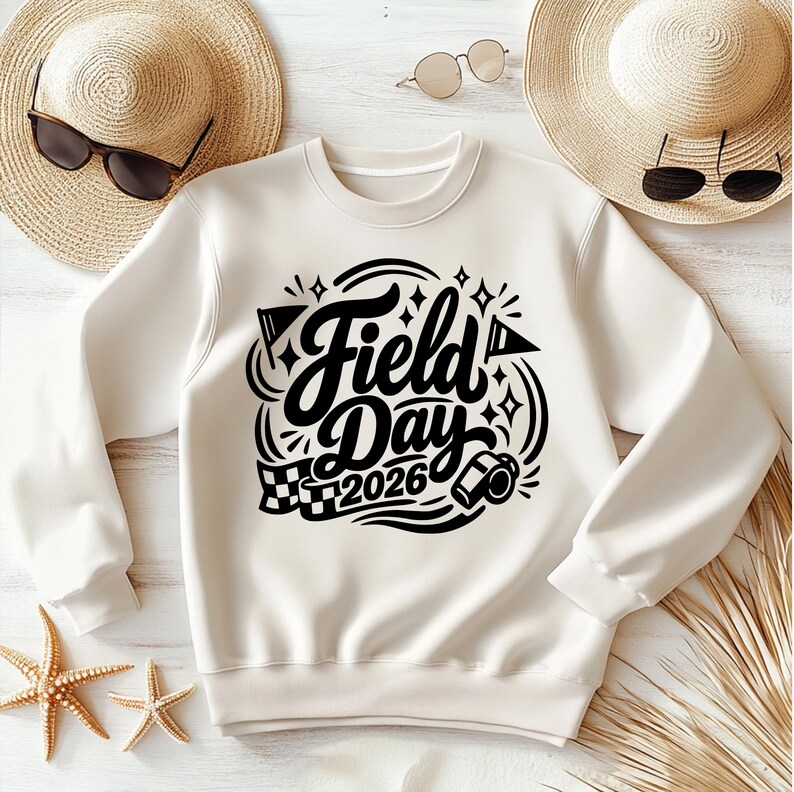 May include: Cream-colored sweatshirt with the words "Field Day 2026" in a black, decorative font. The design includes flags, stars, and a checkered flag. The sweatshirt is laid flat with sunglasses and a straw hat in the background.