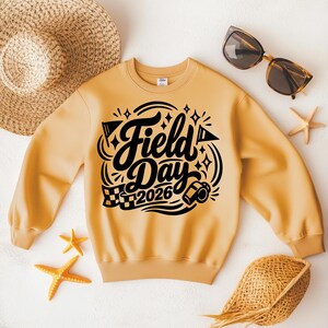 May include: A light orange sweatshirt with the words "Field Day 2026" in black script. The design includes flags, stars, and a checkered flag. The sweatshirt is surrounded by a straw hat, sunglasses, and starfish.