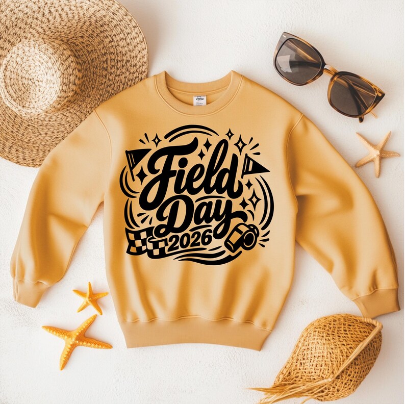 May include: A light orange sweatshirt with the words "Field Day 2026" in black script. The design includes flags, stars, and a checkered flag. The sweatshirt is surrounded by a straw hat, sunglasses, and starfish.