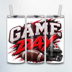 May include: A white cylindrical tumbler with a straw, featuring the text GAME DAY in bold, distressed black and white lettering. The design includes red brushstroke accents, a brown football with white laces, and a black football helmet.