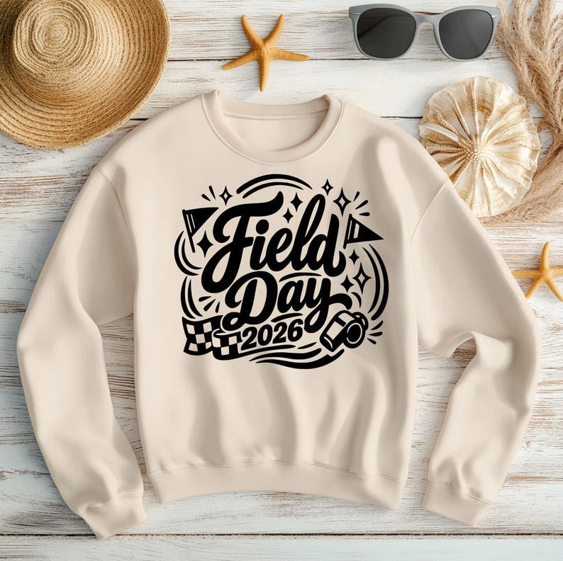 May include: A cream-colored sweatshirt with the words "Field Day 2026" in a black, decorative font. The design includes flags, stars, and a whistle. The sweatshirt is laid on a light wood surface with a straw hat, sunglasses, and seashells.