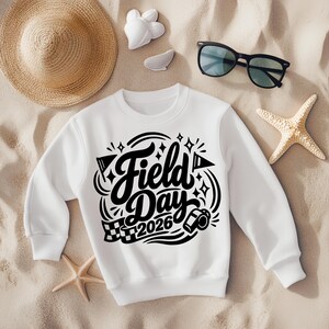 May include: White crewneck sweatshirt with the words "Field Day 2026" in black script. The design includes a whistle, flags, and stars. The sweatshirt is laid on sand with a straw hat, sunglasses, and starfish.