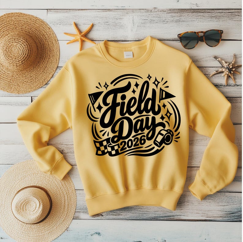 May include: A yellow sweatshirt with the words "Field Day 2026" in black script. The design includes flags, stars, and a checkered flag. The sweatshirt is displayed with a straw hat, sunglasses, and starfish.