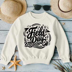 May include: Cream-colored sweatshirt with the words "Field Day 2026" in a black, decorative font. The design includes flags, a checkered flag, and a whistle. The sweatshirt is surrounded by straw hats, sunglasses, and starfish.