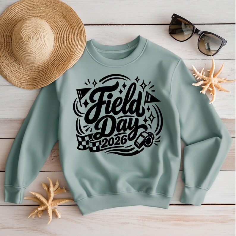May include: A light teal sweatshirt with the words "Field Day 2026" in black script. The design includes stars, flags, and a checkered flag. A straw hat, sunglasses, and starfish are also visible.