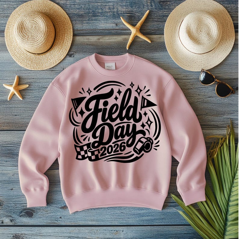 May include: A light pink sweatshirt with the words "Field Day 2026" in a black, stylized font. The design includes flags, diamonds, and a checkered flag. The sweatshirt is surrounded by beach accessories.