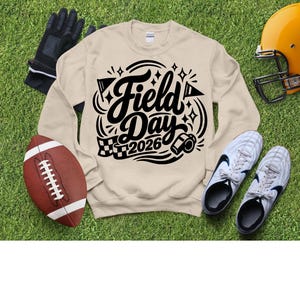 May include: A beige sweatshirt with the words "Field Day 2026" in black script and graphics. The image includes a football, a helmet, cleats, and gloves, all on a green grassy surface.