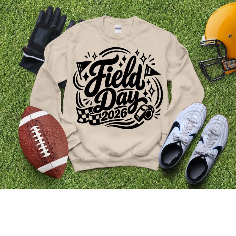May include: A beige sweatshirt with the words "Field Day 2026" in black script and graphics. The image includes a football, a helmet, cleats, and gloves, all on a green grassy surface.