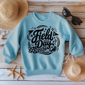 May include: A light blue sweatshirt with the words "Field Day 2026" in black script. The design includes stars, a checkered flag, and a camera. The sweatshirt is displayed with a straw hat, sunglasses, and starfish.