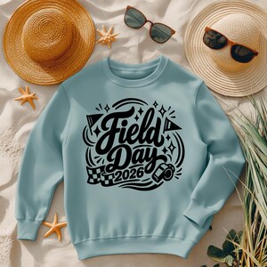 May include: A light blue sweatshirt with the words "Field Day 2026" in a black, decorative font. The design includes flags, stars, and a checkered flag. The sweatshirt is displayed with a straw hat, sunglasses, and starfish.