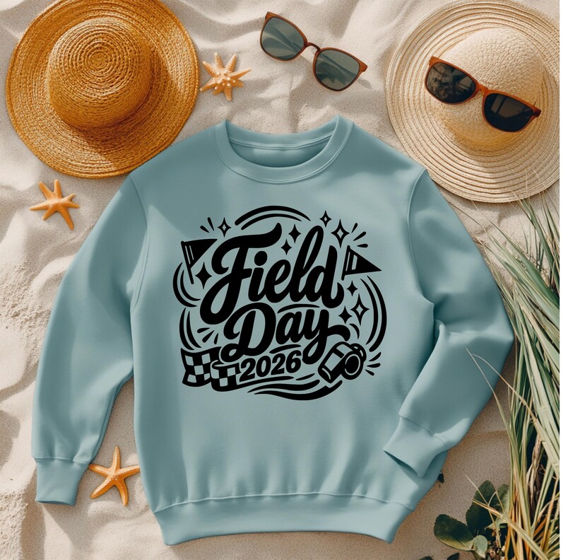 May include: A light blue sweatshirt with the words "Field Day 2026" in a black, decorative font. The design includes flags, stars, and a checkered flag. The sweatshirt is displayed with a straw hat, sunglasses, and starfish.