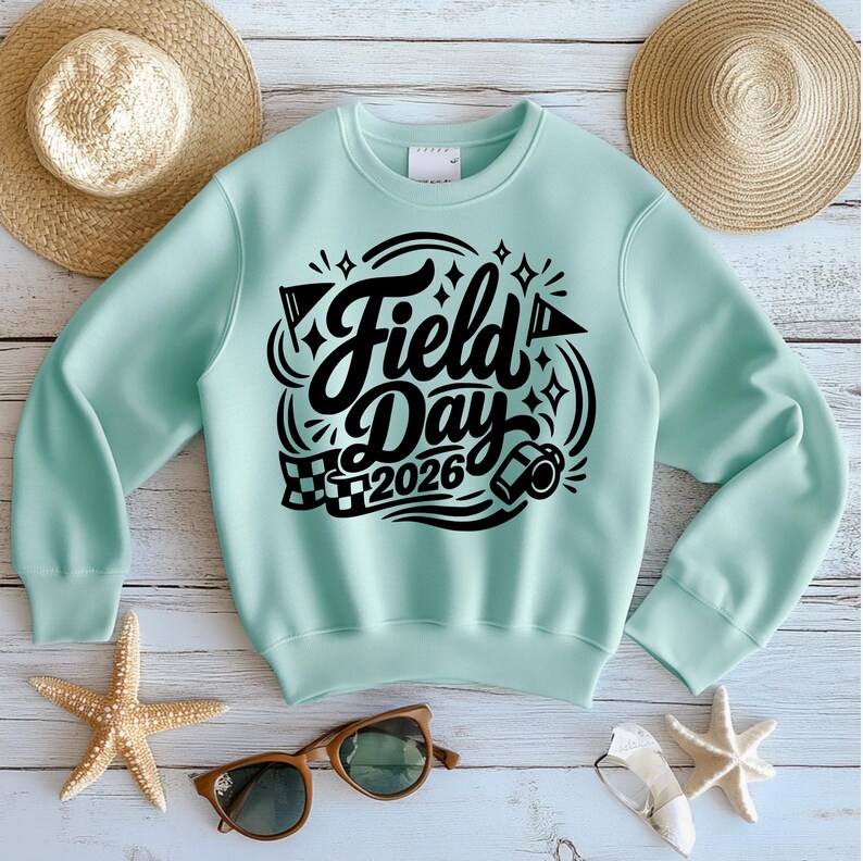 May include: A light teal sweatshirt with the words "Field Day 2026" in a black, stylized font. The design includes stars, flags, and a race car. The sweatshirt is displayed on a light wood-grain surface with a straw hat, sunglasses, and starfish.