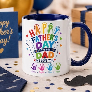 Könnte beinhalten: Weißer Keramikbecher mit blauem Henkel und Rand, mit dem Text "Happy Father's Day" in bunten Buchstaben. Der Becher enthält Handabdrücke und die Namen Emma, Sophia, Liam und Noah. Eine marineblaue Krawatte und Karte sind im Hintergrund.