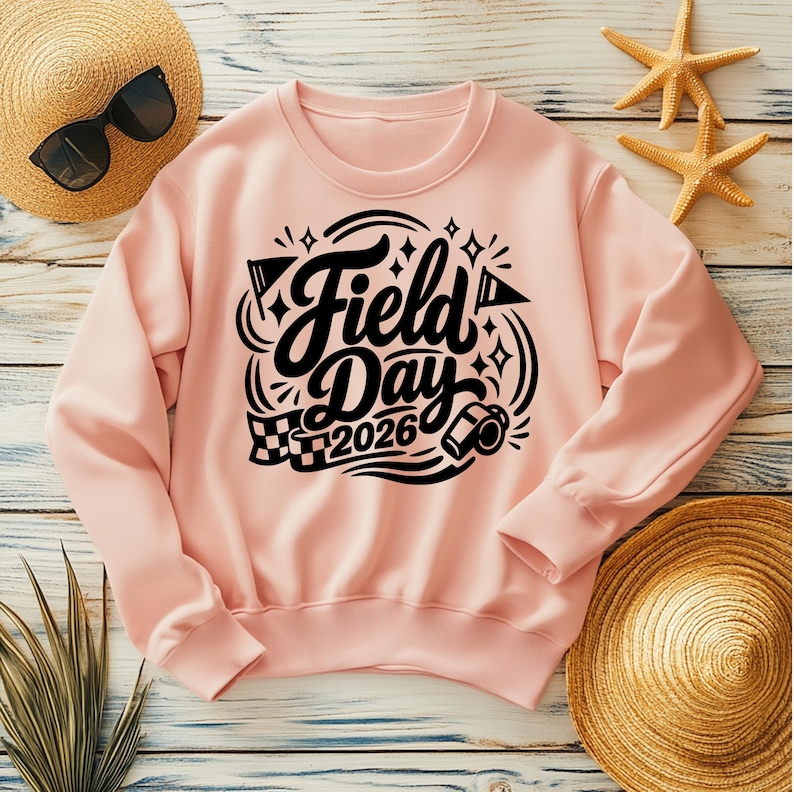 May include: A light pink sweatshirt with the words "Field Day 2026" in a black, decorative font. The design includes a checkered flag, a film reel, and star accents. A straw hat and sunglasses are visible in the background.