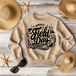 May include: A tan sweatshirt with the words "Field Day 2026" in black script and graphics. The sweatshirt is surrounded by straw hats, sunglasses, and starfish on a light wood background.