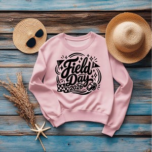 May include: A light pink sweatshirt with the words "Field Day 2026" in a black, decorative font. The design includes a checkered flag, a megaphone, and other graphic elements. The sweatshirt is displayed on a blue and brown wooden surface, with a straw hat and sunglasses.