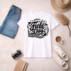 May include: White t-shirt with a black graphic that reads "Field Day 2026". The design includes flags, stars, and a checkered flag. The shirt is laid flat with blue jeans, a camera, a notebook, a hat, shoes, and a cup of coffee.