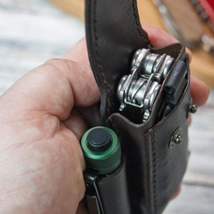 Puede incluir: Una funda de cuero marr&oacute;n para multiherramientas con una multiherramienta y una linterna verde. La funda est&aacute; abierta, revelando las herramientas plateadas. Una navaja roja est&aacute; en el fondo.