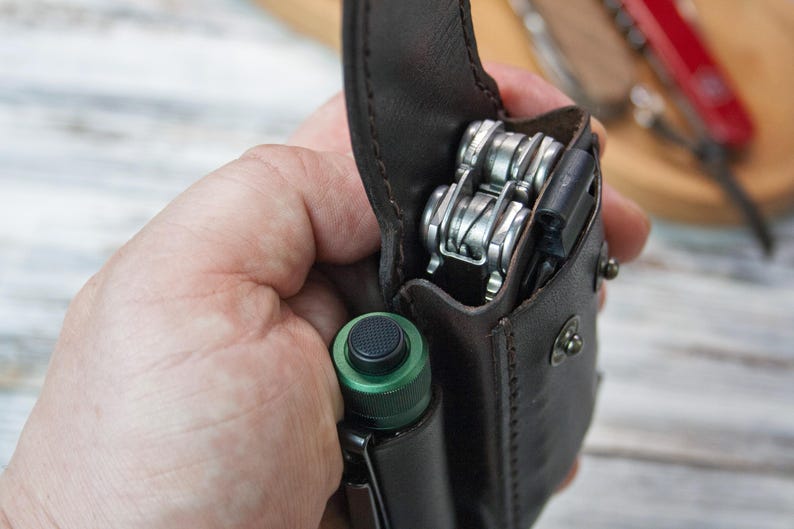 Puede incluir: Una funda de cuero marr&oacute;n para multiherramientas con una multiherramienta y una linterna verde. La funda est&aacute; abierta, revelando las herramientas plateadas. Una navaja roja est&aacute; en el fondo.
