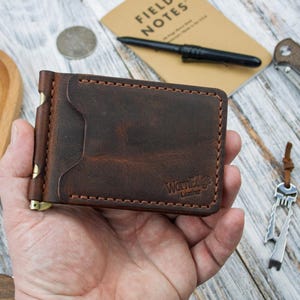 May include: A brown leather wallet with a money clip, featuring the "WornEdge Leather" logo. The wallet is surrounded by a red Swiss Army knife, a small black pen, a folding knife, and a key chain. The items are on a light wood surface.