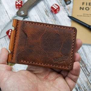 Puede incluir: Una cartera de cuero marrón con un clip para billetes, con una superficie texturizada y bordes cosidos. La cartera se sostiene en una mano, con un cuchillo, dados, un bolígrafo y un cuaderno con las palabras "FIELD NOTES" visibles en el fondo.