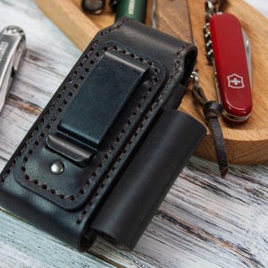 Puede incluir: Una funda de cuero negro para multiherramientas con clip para cintur&oacute;n, junto a una multiherramienta plateada y una navaja suiza roja. La funda tiene un dise&ntilde;o cosido y un peque&ntilde;o bolsillo cil&iacute;ndrico. Los art&iacute;culos est&aacute;n sobre una superficie de madera.