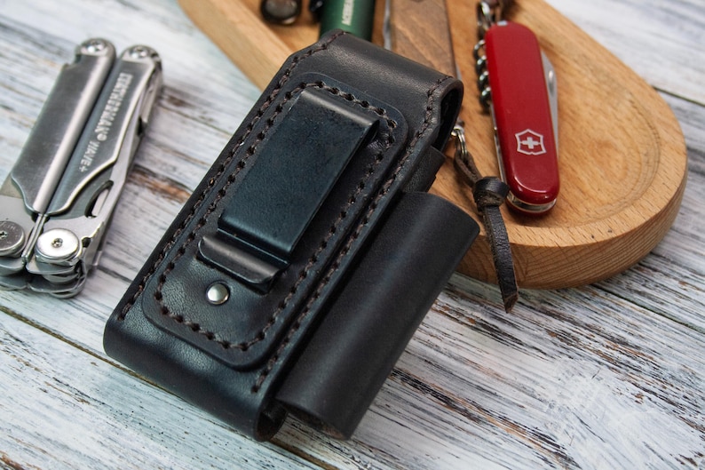 Puede incluir: Una funda de cuero negro para multiherramientas con clip para cintur&oacute;n, junto a una multiherramienta plateada y una navaja suiza roja. La funda tiene un dise&ntilde;o cosido y un peque&ntilde;o bolsillo cil&iacute;ndrico. Los art&iacute;culos est&aacute;n sobre una superficie de madera.