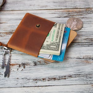 May include: A brown leather wallet with a snap closure, holding US dollar bills and credit cards. A silver coin and a multi-tool are next to the wallet. A wooden Swiss Army knife is attached to the wallet with a leather strap. The items are on a white wooden surface.