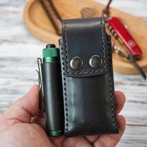 Puede incluir: Un estuche de cuero negro para linterna con dos cierres de bot&oacute;n a presi&oacute;n plateados y un estuche de linterna de cuero negro a juego. Una linterna verde y negra est&aacute; en un soporte de clip de cuero negro. Una navaja suiza roja est&aacute; en el fondo.