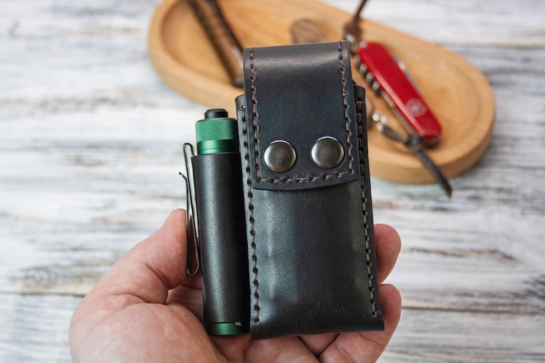 Puede incluir: Un estuche de cuero negro para linterna con dos cierres de bot&oacute;n a presi&oacute;n plateados y un estuche de linterna de cuero negro a juego. Una linterna verde y negra est&aacute; en un soporte de clip de cuero negro. Una navaja suiza roja est&aacute; en el fondo.